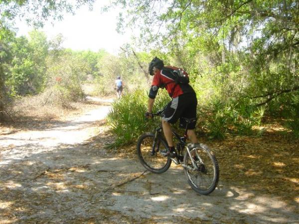 A cyclist riding a mountain bike along a dirt trail in a wooded area, surrounded by greenery and scattered leaves. Another cyclist can be seen in the background, navigating the path. The scene is bright and sunny, indicating a warm day. Alafia River State Park mountain bike trail.