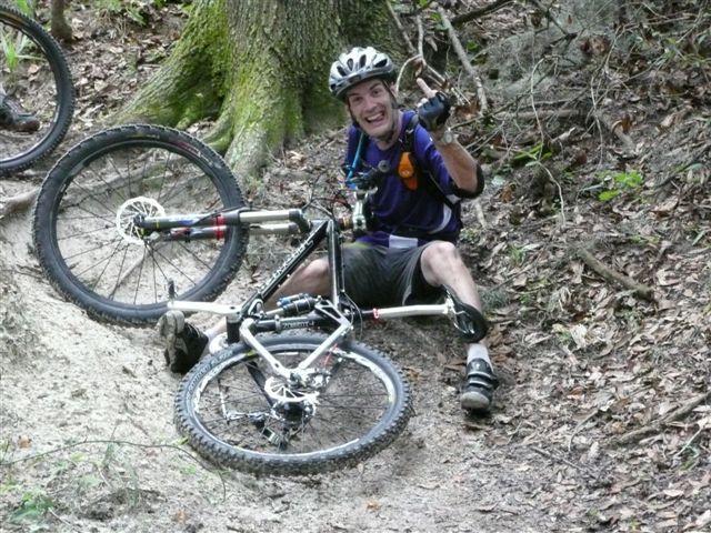 A mountain biker sitting on the ground next to a fallen bike, smiling and giving a thumbs-up. The scene is set in a wooded area with a mix of dirt and leaves on the ground. Alafia River State Park mountain bike trail.