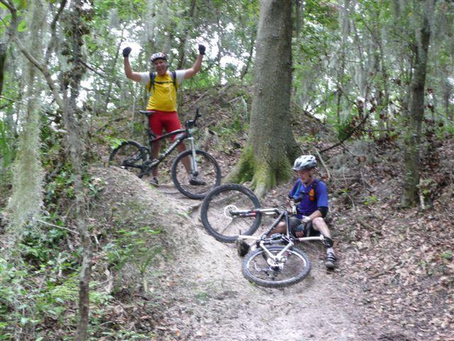 Two mountain bikers are enjoying a trail in a wooded area. One rider, wearing a yellow shirt and red shorts, stands triumphantly with raised arms at the top of a hill, while the other, dressed in a blue shirt, sits beside his bike on a sandy path. Surrounding them are trees and greenery, typical of a natural trail environment. Alafia River State Park mountain bike trail.