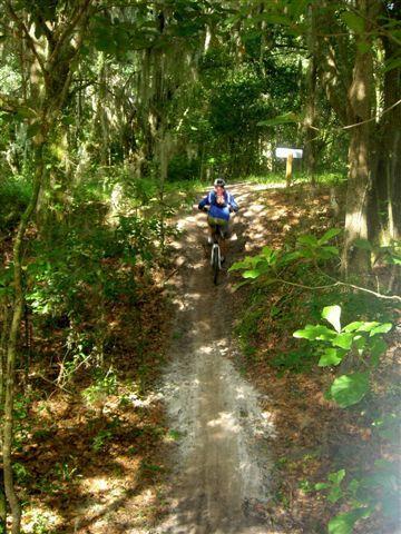 A person riding a mountain bike along a dirt trail in a wooded area, surrounded by trees and lush vegetation. The trail is slightly inclined and there is a sign visible in the background. Sunlight filters through the leaves, creating a dappled light effect on the ground. Alafia River State Park mountain bike trail.