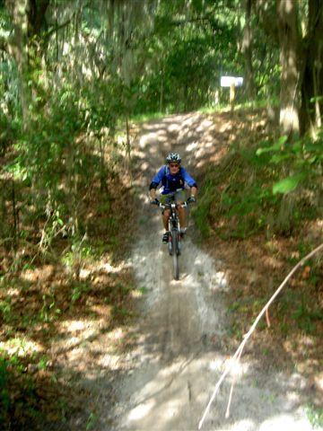 A mountain biker wearing a helmet and blue cycling gear rides down a dirt trail surrounded by lush green foliage and trees. The path is lined with fallen leaves, and there is a signpost visible in the background. Alafia River State Park mountain bike trail.