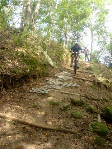 A mountain biker navigating a rocky trail with scattered metal discs on the ground, surrounded by trees and natural foliage. Another cyclist is visible in the background, riding up the hill. Alafia River State Park mountain bike trail.