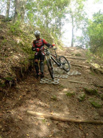 A mountain biker standing beside a bicycle on a steep, rugged trail surrounded by trees. The trail features wooden steps and rocky terrain, showcasing a natural outdoor environment suitable for mountain biking. Alafia River State Park mountain bike trail.