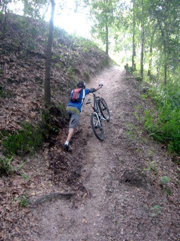 A person walking uphill on a dirt trail while carrying a mountain bike on their shoulder. The path is lined with trees and greenery, indicating a natural outdoor setting. Alafia River State Park mountain bike trail.