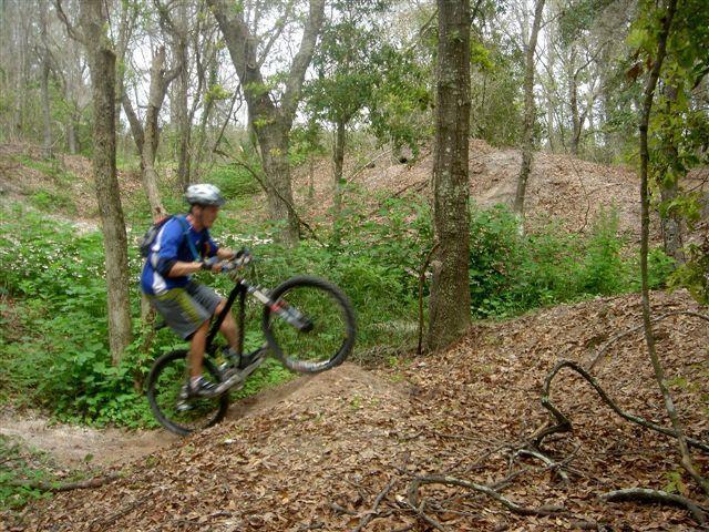 A mountain biker performing a wheelie on a dirt path surrounded by trees and greenery, with fallen leaves scattered on the ground. Alafia River State Park mountain bike trail.