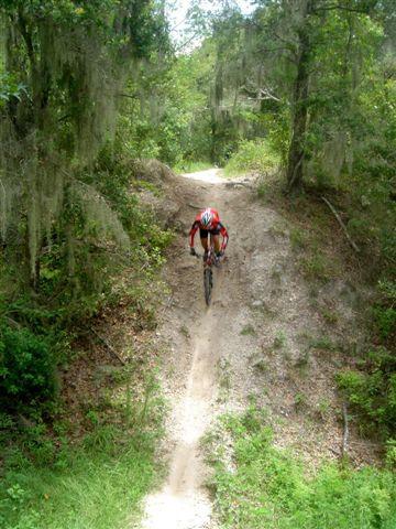 A mountain biker in red gear descends a rugged trail surrounded by lush greenery and trees, showcasing an action shot of jumping off a dirt incline. Alafia River State Park mountain bike trail.