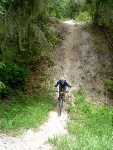 A mountain biker riding up a dirt trail with greenery and trees on either side, showcasing a steep incline. The cyclist is wearing a helmet and blue clothing. Alafia River State Park mountain bike trail.