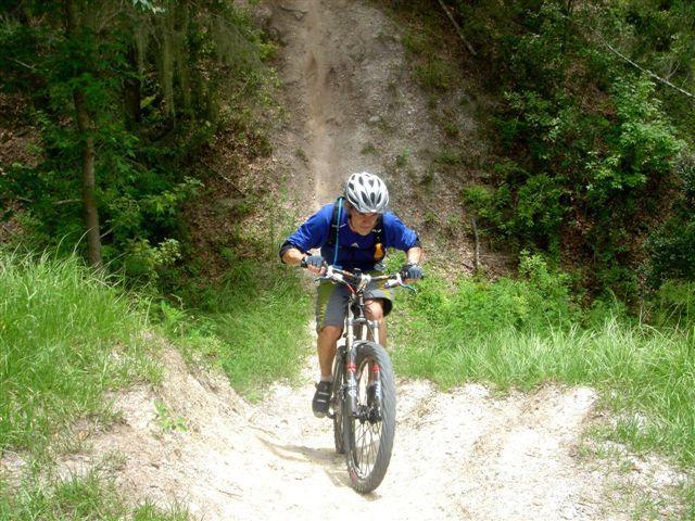 A person wearing a helmet and a blue shirt is riding a mountain bike up a sandy trail, surrounded by green grass and trees. The cyclist appears focused as they navigate the incline. Alafia River State Park mountain bike trail.