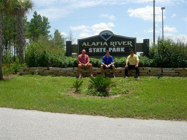 Three individuals sitting on a stone wall in front of the Alafia River State Park sign. The scene features green grass, shrubs, and palm trees under a bright blue sky with fluffy white clouds. The men are casually dressed, enjoying a sunny day at the park. Alafia River State Park mountain bike trail.