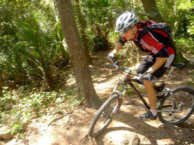 A mountain biker navigating a dirt trail through a wooded area, wearing a helmet and sporty attire, with trees and foliage in the background. Alafia River State Park mountain bike trail.