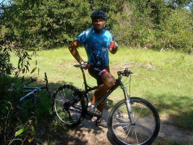 A cyclist in a blue shirt and black shorts stands beside a mountain bike on a grassy trail, smiling and holding a beverage. Sunlight filters through the trees in the background, creating a vibrant outdoor setting. Alafia River State Park mountain bike trail.
