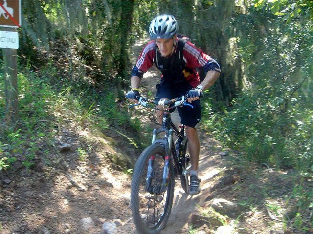 A mountain biker riding on a rocky forest trail, wearing a helmet and a red and black jersey, with trees and greenery in the background. Alafia River State Park mountain bike trail.