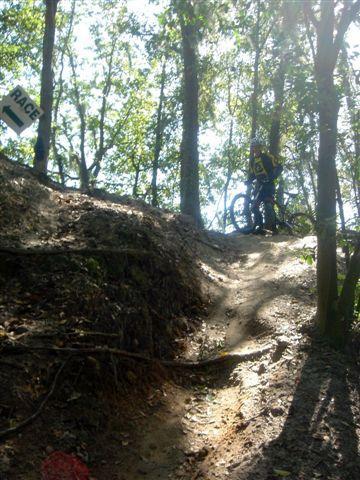 A mountain biker navigating a dirt trail in a forested area, with sunlight filtering through the trees. A race sign is visible in the background, indicating an off-road biking event. The trail is winding and shows various natural features like roots and uneven terrain. Alafia River State Park mountain bike trail.