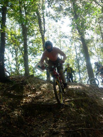 A mountain biker wearing a helmet and no shirt descends a steep, wooded trail, surrounded by trees and sunlight filtering through the leaves. Other bikers are visible in the background. Alafia River State Park mountain bike trail.