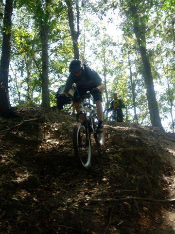 A mountain biker descending a rocky slope in a forested area, with sunlight filtering through the trees. Another cyclist can be seen in the background, slightly out of focus. Alafia River State Park mountain bike trail.