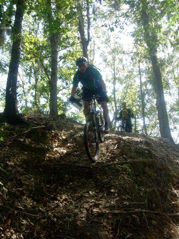 A mountain biker navigating a steep descent on a dirt trail surrounded by trees, with another cyclist visible in the background. The sunlight filters through the foliage, creating a dynamic and adventurous atmosphere. Alafia River State Park mountain bike trail.