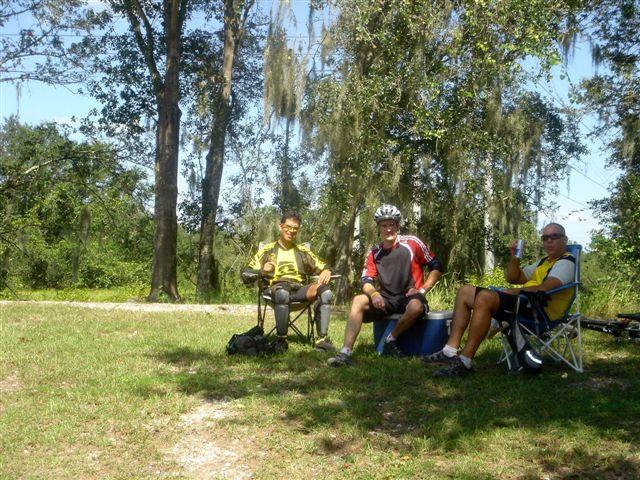 Three cyclists take a break outdoors, sitting in lawn chairs under the shade of trees. One person wears a yellow jersey and protective gear, while another is in a red and black jersey with a cycling helmet. The third individual wears a yellow shirt and holds a drink, enjoying the sunny day in a grassy area. Alafia River State Park mountain bike trail.