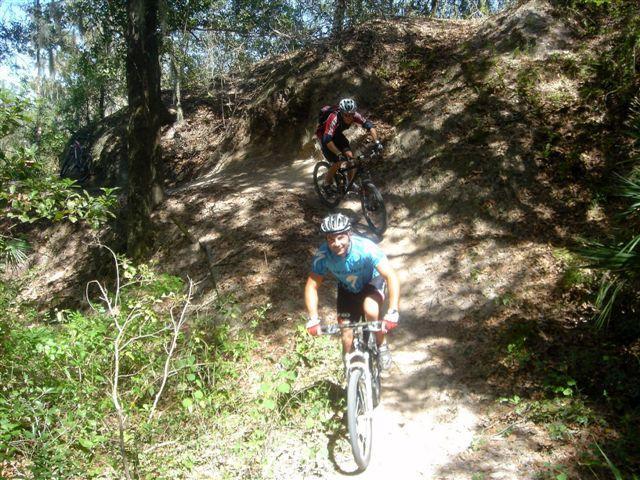 Two mountain bikers navigating a dirt trail surrounded by lush greenery. One rider, in a blue shirt, is in the foreground smiling as they pedal uphill, while another rider, in a red shirt, is positioned further up the trail. The scene captures a sunny day with dappled sunlight filtering through the trees. Alafia River State Park mountain bike trail.