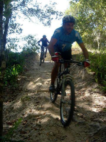 Two mountain bikers navigate a sandy trail surrounded by trees and greenery. One rider, in a blue jersey and helmet, is in the foreground, while another rider follows behind in a darker outfit. The sunlight filters through the leaves, creating a vibrant outdoor scene. Alafia River State Park mountain bike trail.