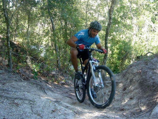 A mountain biker in a blue shirt and helmet rides uphill on a dirt trail surrounded by trees. The cyclist is captured mid-jump, showcasing an active outdoor scene. Alafia River State Park mountain bike trail.