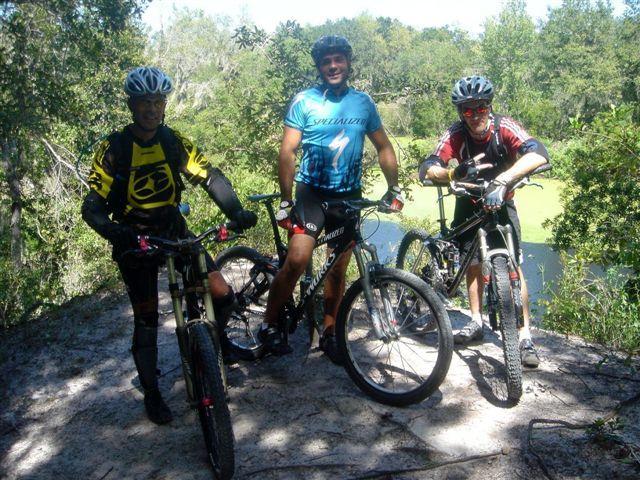 Three mountain bikers posing with their bikes in a wooded area. They are wearing protective gear and colorful cycling jerseys. Behind them, a serene body of water is partially visible, surrounded by trees. The scene reflects an outdoor adventure environment. Alafia River State Park mountain bike trail.