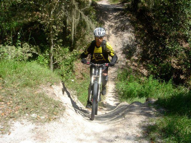 A mountain biker wearing a helmet and protective gear rides up a sandy incline on a trail surrounded by lush greenery and trees. The path is winding with a mix of dirt and grass, indicating a natural outdoor setting. Alafia River State Park mountain bike trail.