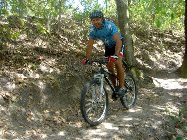 A man riding a mountain bike on a dirt trail surrounded by trees and foliage, wearing a blue cycling jersey and shorts, smiling as he navigates the path. Alafia River State Park mountain bike trail.