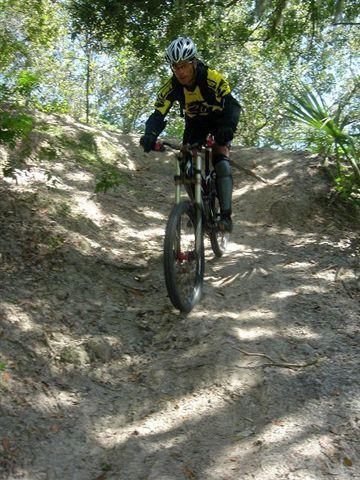 A person riding a mountain bike down a sandy, uneven trail surrounded by trees. The cyclist is wearing a helmet and protective gear, with sunlight filtering through the foliage above. Alafia River State Park mountain bike trail.