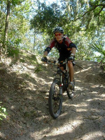 A person riding a mountain bike up a sandy trail surrounded by trees. The cyclist is wearing a helmet, sunglasses, and athletic clothing, with a determined expression on their face. Sunlight filters through the foliage above, creating a lively outdoor atmosphere. Alafia River State Park mountain bike trail.