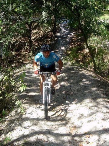 A cyclist in a blue shirt and helmet is riding uphill on a sandy trail surrounded by trees and foliage. Alafia River State Park mountain bike trail.