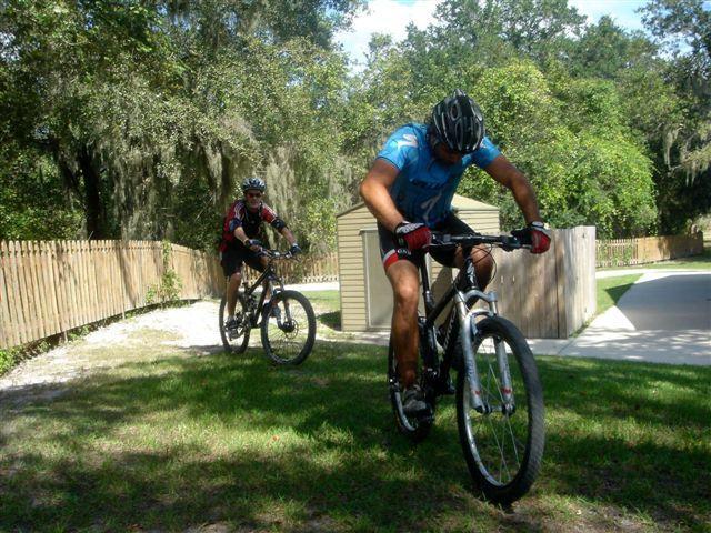Two mountain bikers ride on a grassy path surrounded by trees and a wooden fence. One cyclist, wearing a blue shirt and helmet, is in the foreground, while another, clad in a red and black outfit, follows closely behind. A storage shed is visible in the background, set against a backdrop of greenery. Alafia River State Park mountain bike trail.