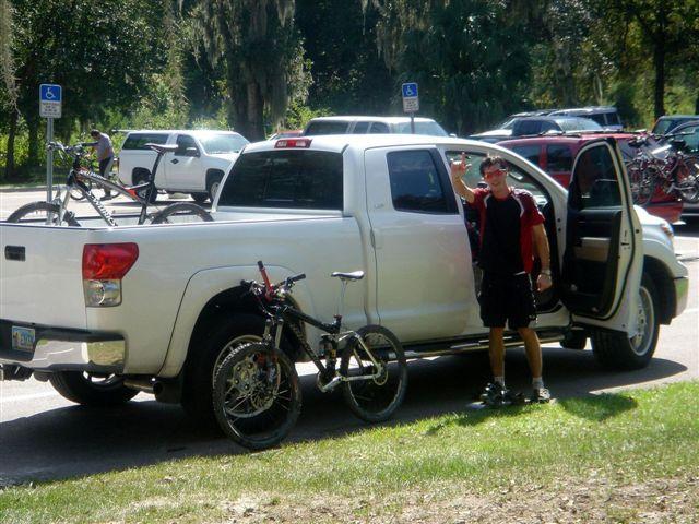 A man stands next to a white pickup truck parked on a sunny day, waving and smiling. The truck has bicycles securely mounted in the bed, and another bicycle is parked beside it. The scene is set in a parking area surrounded by greenery, with several vehicles visible in the background. Alafia River State Park mountain bike trail.