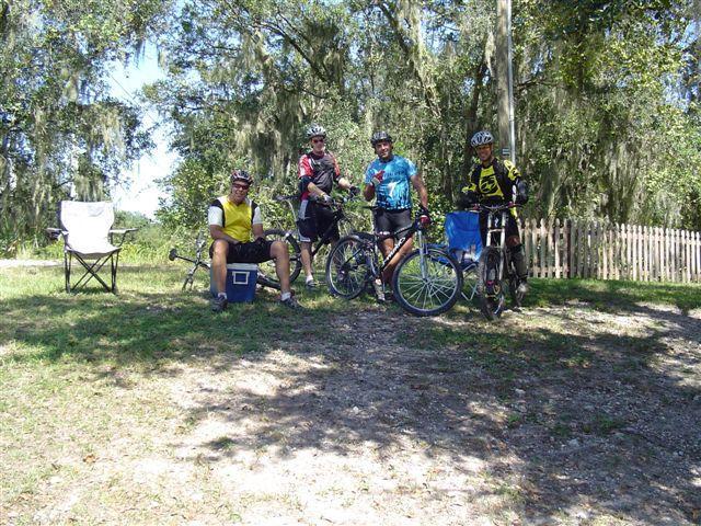 A group of four cyclists relaxing outdoors, sitting beside their bicycles. They are positioned on a grassy area with a few lawn chairs and a cooler nearby. The background features trees and a wooden fence, suggesting a rural or park-like setting. The cyclists are wearing helmets and colorful biking apparel, enjoying a break during their ride. Alafia River State Park mountain bike trail.