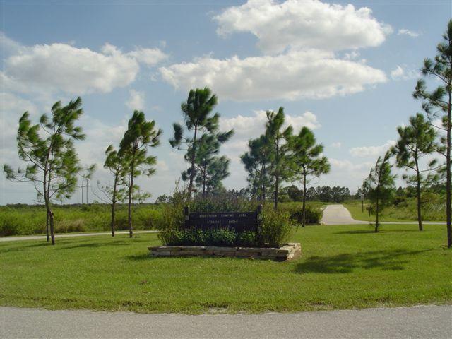 Entryway to a scenic park with a sign surrounded by greenery and small trees, set against a backdrop of a blue sky with fluffy clouds. A winding road leads into the distance. Alafia River State Park mountain bike trail.