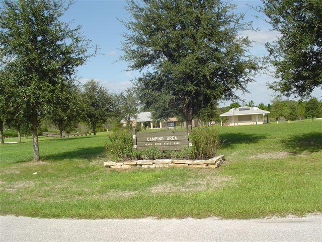 A sign reading "Camping Area" surrounded by green grass and trees, indicating a designated camping site within a park. In the background, there are several structures, likely facilities for campers, under a clear blue sky. Alafia River State Park mountain bike trail.