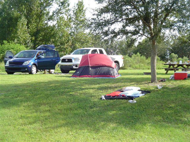 A scenic camping site featuring a red and black tent set up on green grass. Two parked cars, one blue and one white, are visible nearby. A picnic table and cooler can be seen in the background, surrounded by trees and foliage, creating a peaceful outdoor atmosphere. Clothing is spread out on the ground, indicating preparation for camping activities. Alafia River State Park mountain bike trail.