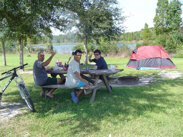 A group of three people sitting at a picnic table outside, enjoying a meal during a camping trip. They are smiling and waving at the camera. A bicycle is leaning against the table, and a colorful tent is set up nearby, with a scenic view of a lake and trees in the background. Alafia River State Park mountain bike trail.
