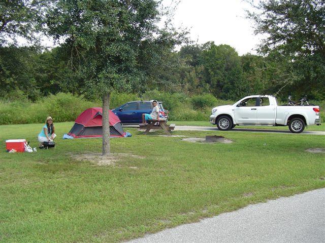 A campsite scene featuring a red tent, a picnic table, and a white pickup truck parked nearby. Two people are present: one is sitting at the picnic table, and the other is preparing food near the tent. Lush greenery and trees are visible in the background, with a peaceful outdoor setting. Alafia River State Park mountain bike trail.