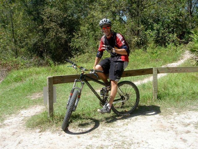 A mountain biker wearing a helmet and sunglasses sits on a black bicycle near a wooden fence, giving a thumbs-up gesture. The background features lush greenery and a dirt path, suggesting a sunny outdoor trail. Alafia River State Park mountain bike trail.