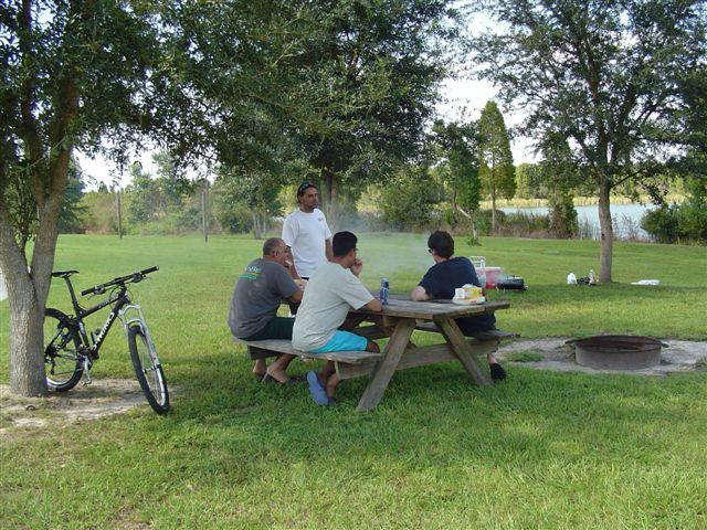 A group of four people seated at a wooden picnic table under a tree in a grassy area, chatting and enjoying food. A person stands at the head of the table. In the background, there is a bicycle parked nearby, and a fire pit can be seen on the ground. The scene is set near a body of water, surrounded by trees on a sunny day. Alafia River State Park mountain bike trail.