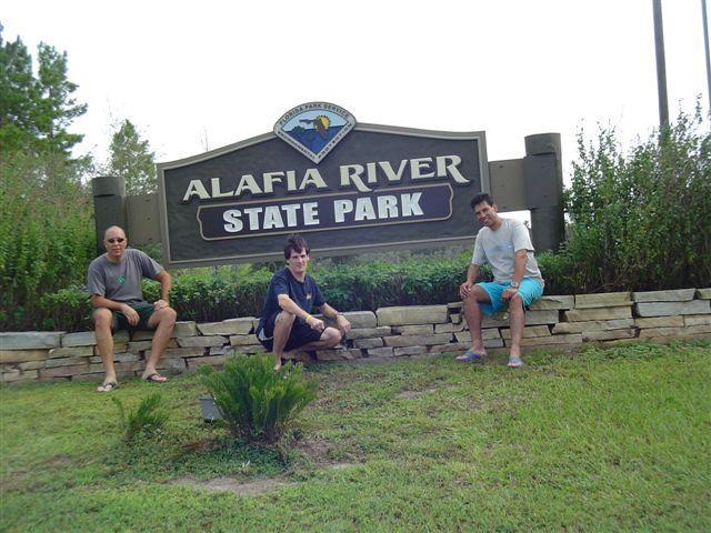 Three individuals sitting on a stone wall in front of a large sign that reads "Alafia River State Park." Surrounding greenery and a grassy area are visible, suggesting a natural outdoor setting. The sky is partly cloudy. Alafia River State Park mountain bike trail.