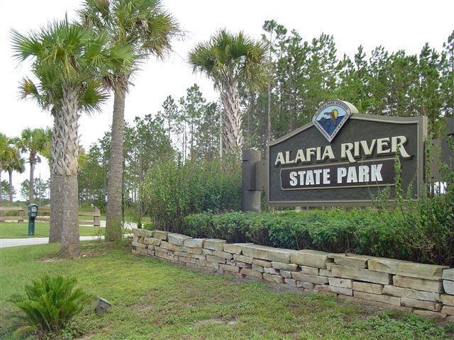 Sign for Alafia River State Park surrounded by palm trees and greenery, with a stone border and a clear blue sky in the background. Alafia River State Park mountain bike trail.