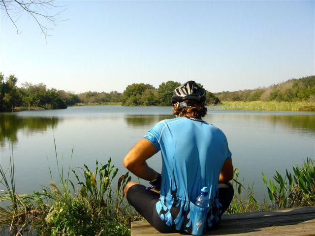 A cyclist wearing a blue jersey sits on a wooden dock, looking out over a calm lake surrounded by greenery. The scene features clear blue skies and distant trees, creating a peaceful outdoor atmosphere. Alafia River State Park mountain bike trail.