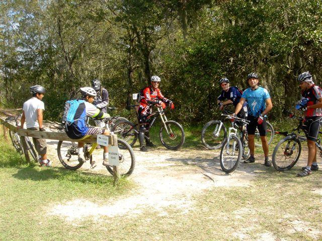 A group of six mountain bikers taking a break at a trail junction, surrounded by trees and greenery. Some riders are seated on a wooden bench, while others stand near their bicycles, dressed in cycling gear and helmets. Alafia River State Park mountain bike trail.