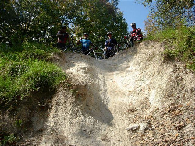 Four mountain bikers standing at the edge of a steep dirt slope, surrounded by greenery and autumn foliage. The slope is sandy, indicating a downhill biking trail. The riders are equipped with helmets and bicycles, preparing for the descent. Alafia River State Park mountain bike trail.