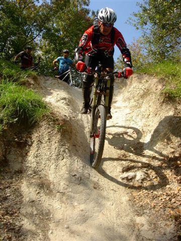 A mountain biker in a red and black outfit navigates a downhill dirt trail, showcasing skill and balance. In the background, fellow cyclists observe the ride under a clear blue sky surrounded by trees. Alafia River State Park mountain bike trail.