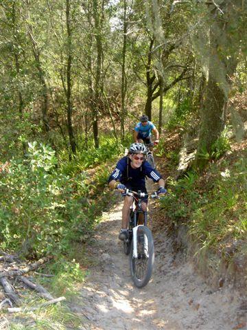 Two mountain bikers navigating a narrow dirt trail surrounded by lush greenery and trees. The first rider is focused and slightly leaning forward, wearing a helmet and blue athletic gear, while the second rider follows closely behind. Sunlight filters through the trees, creating a vibrant outdoor scene. Alafia River State Park mountain bike trail.