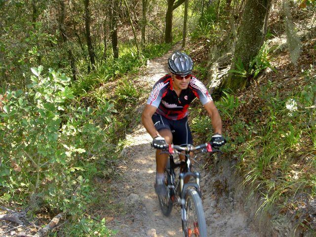 A mountain biker navigating a narrow dirt trail surrounded by trees and foliage, wearing a helmet and cycling gear in shades of red and black. Alafia River State Park mountain bike trail.