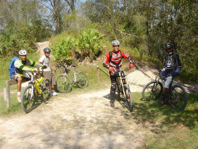 Four mountain bikers gathered at a dirt trail intersection, surrounded by trees and greenery. Two riders are standing next to their bikes, while the other two are seated nearby. One rider wears a bright red and black outfit, while another sports a green and white jersey. The scene captures a sunny day in an outdoor biking environment. Alafia River State Park mountain bike trail.
