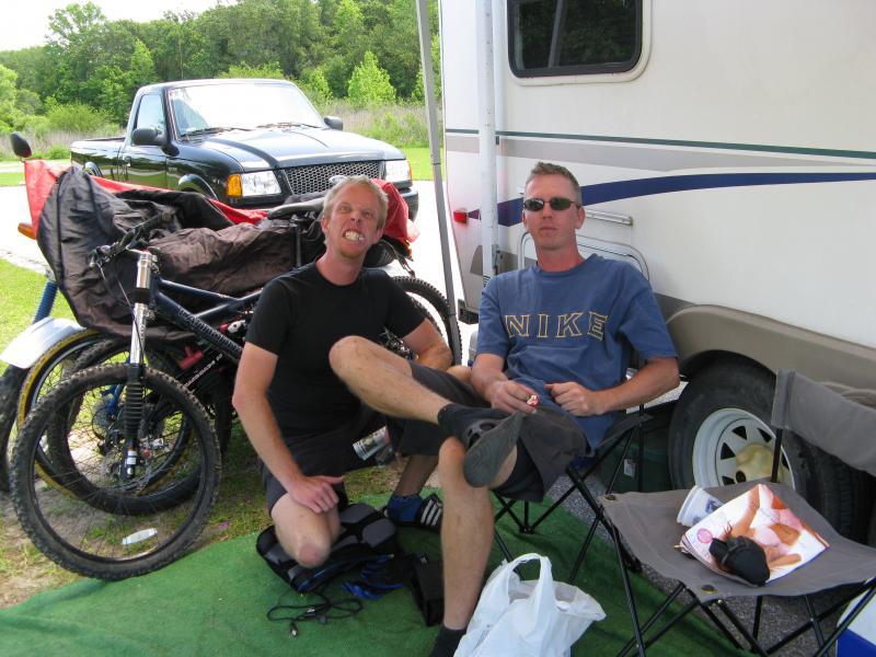 Two men sitting outside a camper, one with short blond hair and a playful expression, and the other with short brown hair wearing sunglasses. They are surrounded by camping gear and bicycles. A black pickup truck is parked in the background, and the setting is a green outdoor area. Alafia River State Park mountain bike trail.